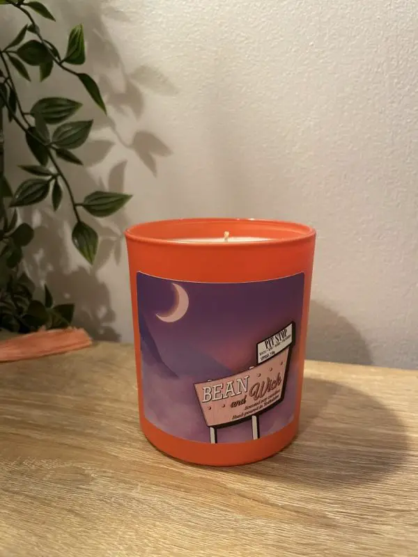 Beanandwick candle. Coral jar with a purple Label. On the label is a Beanandwick motel sign with a purple dreamy backdrop of mountains and a crescent moon.